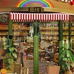 Kahvilan tai leipomon ulkokuva, jossa on punavalkoraidallinen markiisi ja sateenkaari sekä edessä olevia pöytiä ja tuoleja